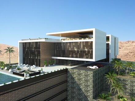 INJ Architects منظور جوي لمجمع سكني هندسي أبيض من طابقين يقع على تضاريس صحراوية صخرية يضم مسبحاً وجدران استنادية من الجابيون ونخيلاً على موقع منحدر