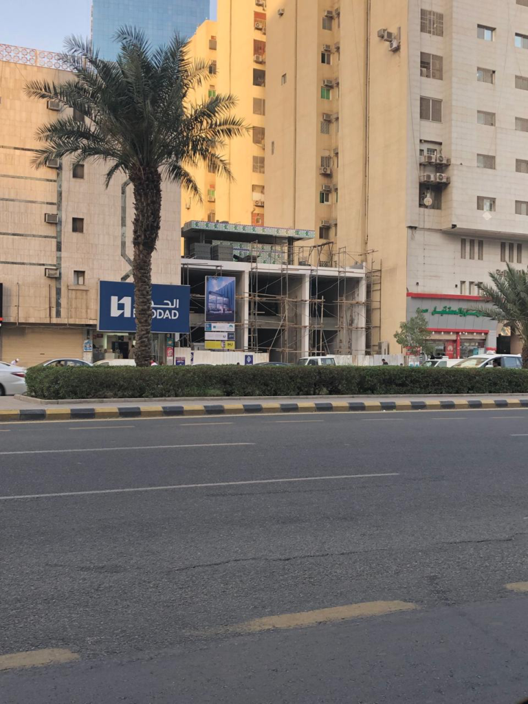 INJ Architects daytime construction photograph showing a multi-level concrete frame building covered in metal scaffolding situated between taller residential buildings on Al-Aziziah Street