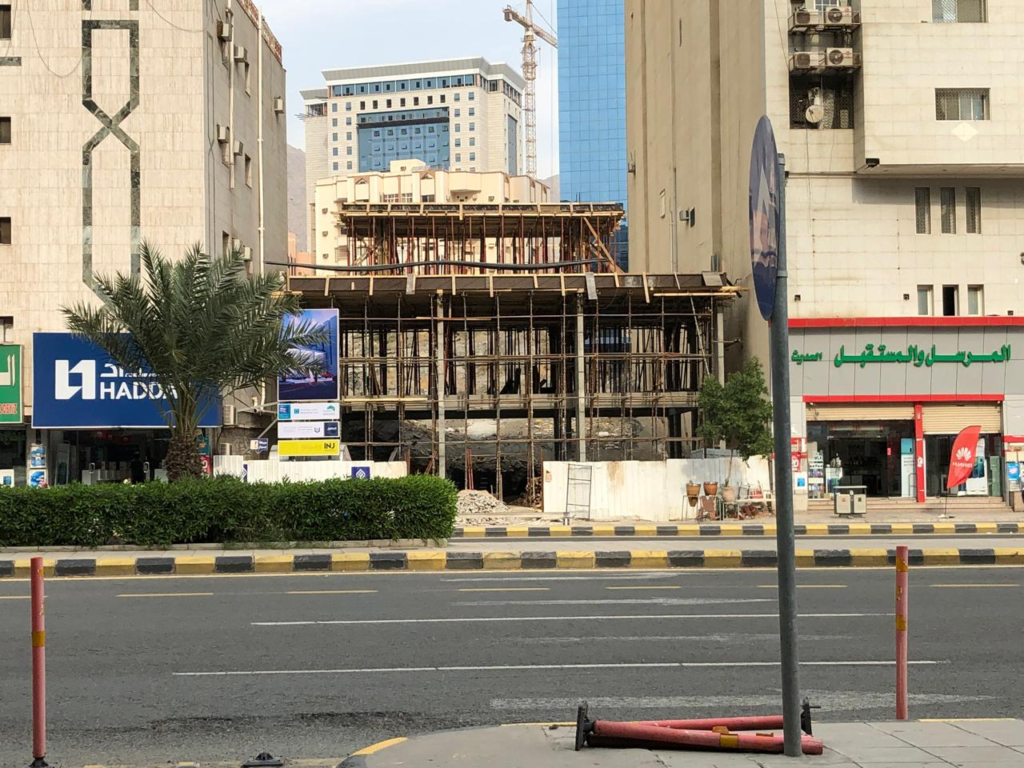 INJ Architects daytime construction photograph showing the exposed concrete column and slab framework of a low-rise commercial building under construction between existing mid-rise structures on a busy street