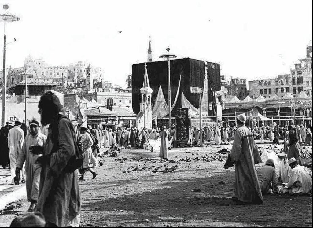 INJ Architects historical black and white photograph capturing pilgrims gathered around the cubic Kaaba structure under a bright sky with surrounding low buildings