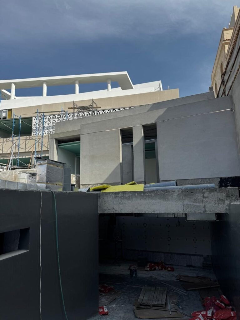 INJ Architects exterior construction site photograph showing raw grey concrete walls in the foreground beneath a completed white upper terrace featuring marble columns and geometric screens