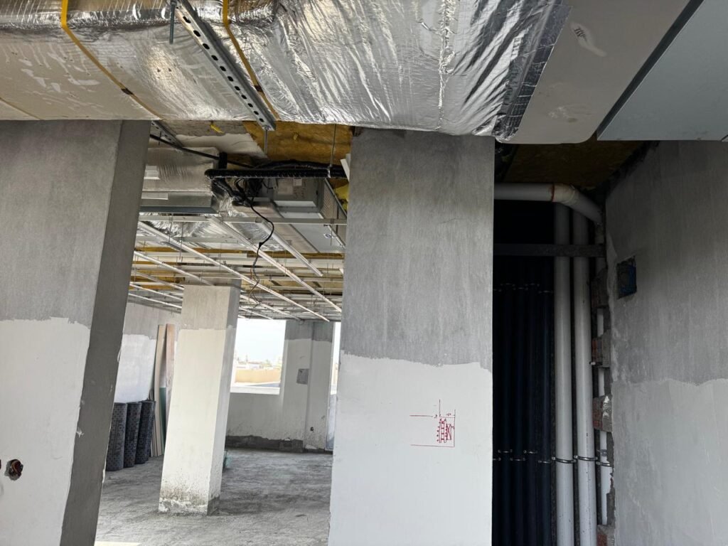 INJ Architects interior construction shot revealing exposed ceiling ductwork insulation and piping above raw concrete columns partially coated in white primer with red architectural markings