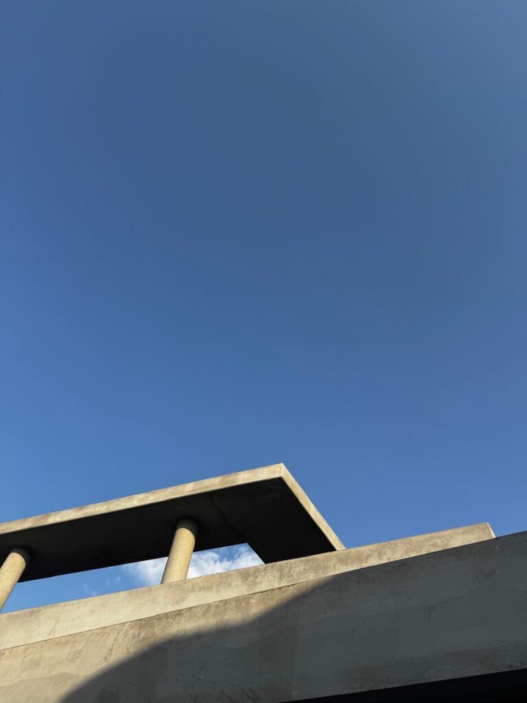 INJ Architects upward perspective photograph of an unfinished grey concrete cantilevered roof plane supported by two cylindrical columns against a vast clear blue sky
