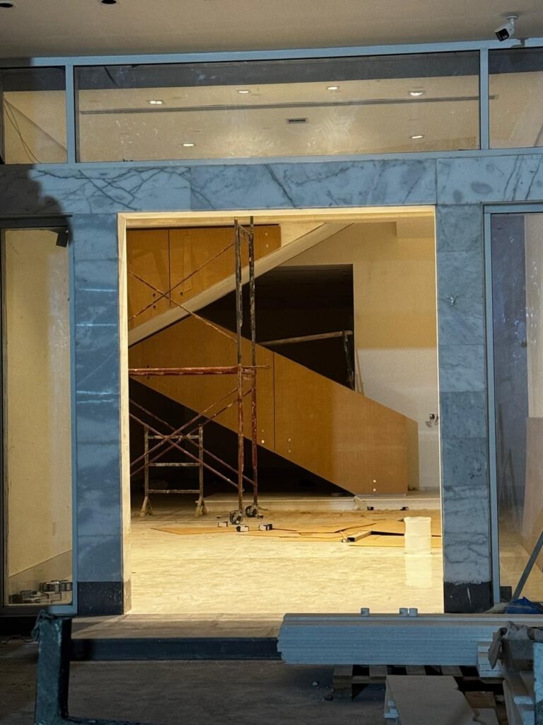 INJ Architects view through a thick marble clad doorway into an unfinished interior space featuring a solid wooden staircase under construction surrounded by red metal scaffolding