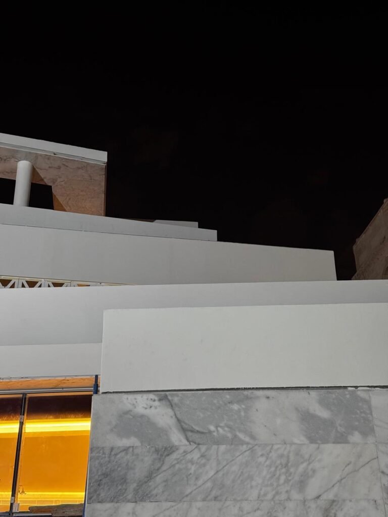 INJ Architects night construction photograph looking upward at stepped white architectural volumes resting on a heavily veined grey marble base with warm interior light escaping below