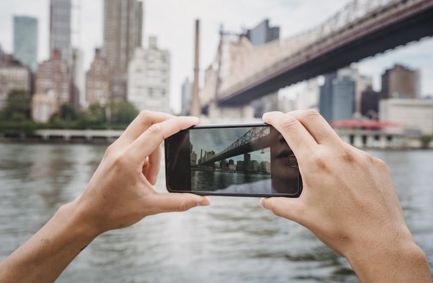 Crop unrecognizable traveler taking photo of modern suspension bridge in city using mobile phone in daytime