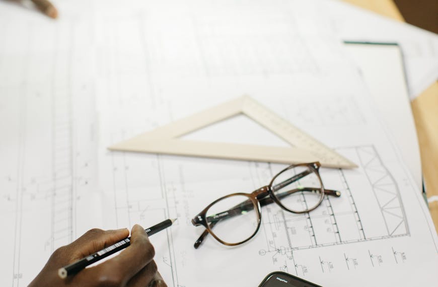 An architect working on blueprints in a modern office with a smartphone and glasses on the desk.