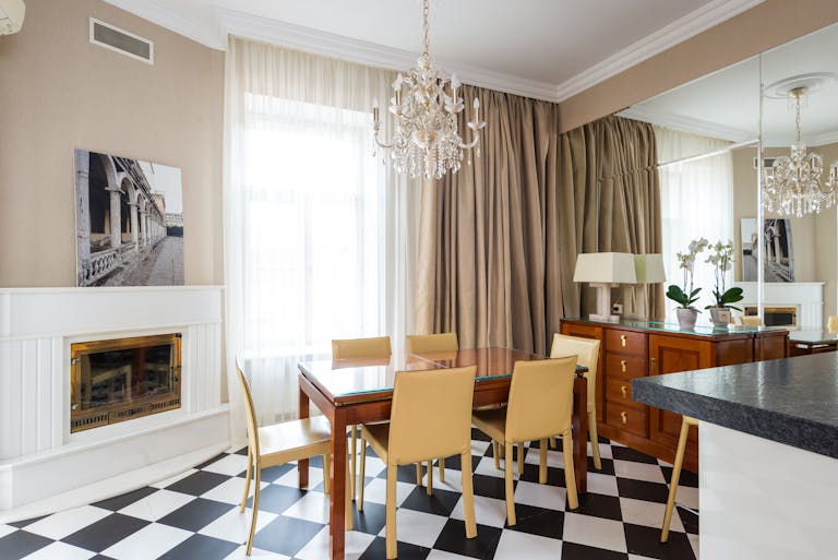 Dining table with chairs placed near window covered with curtains in kitchen with classic chandelier in apartment