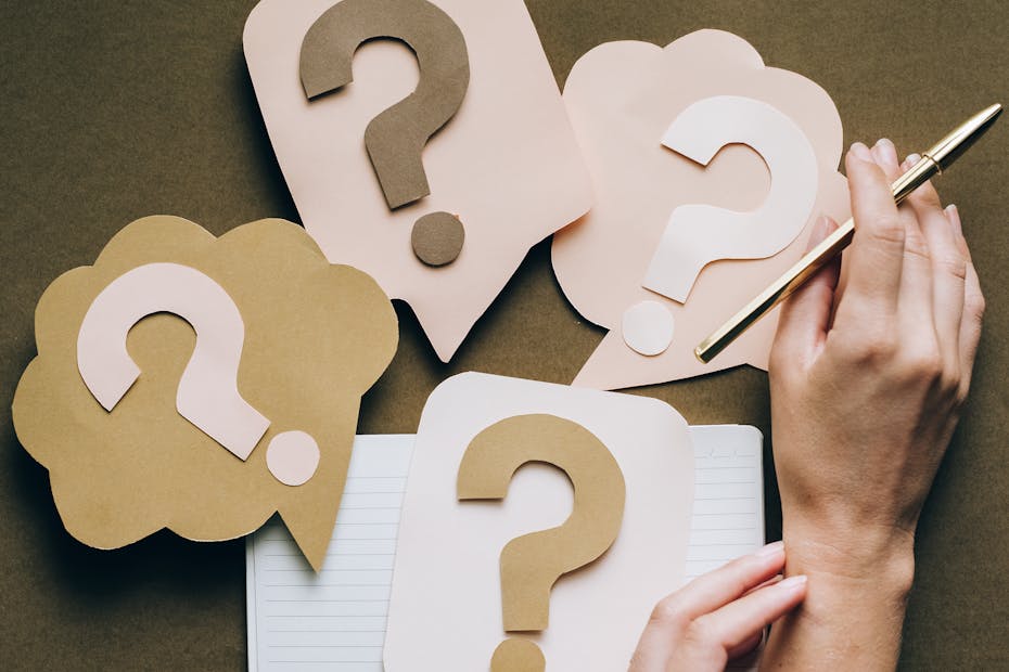 Hands holding pen next to paper question marks on notebook, symbolizing inquiry and creativity.
