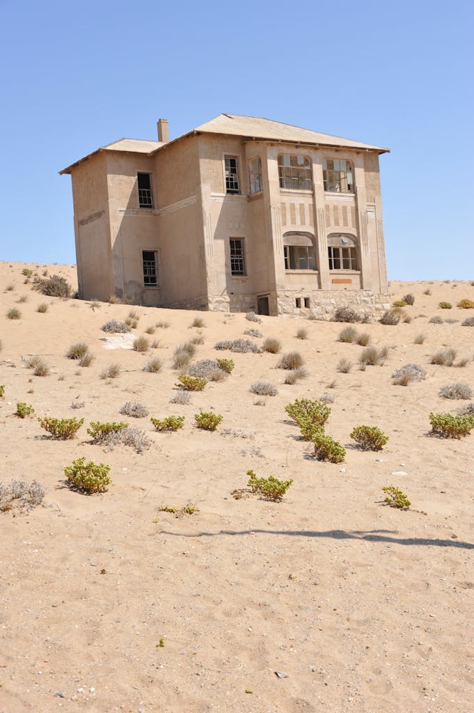 An abandoned house in Kolmanskop, Namibia, surrounded by desert sand and sparse vegetation.