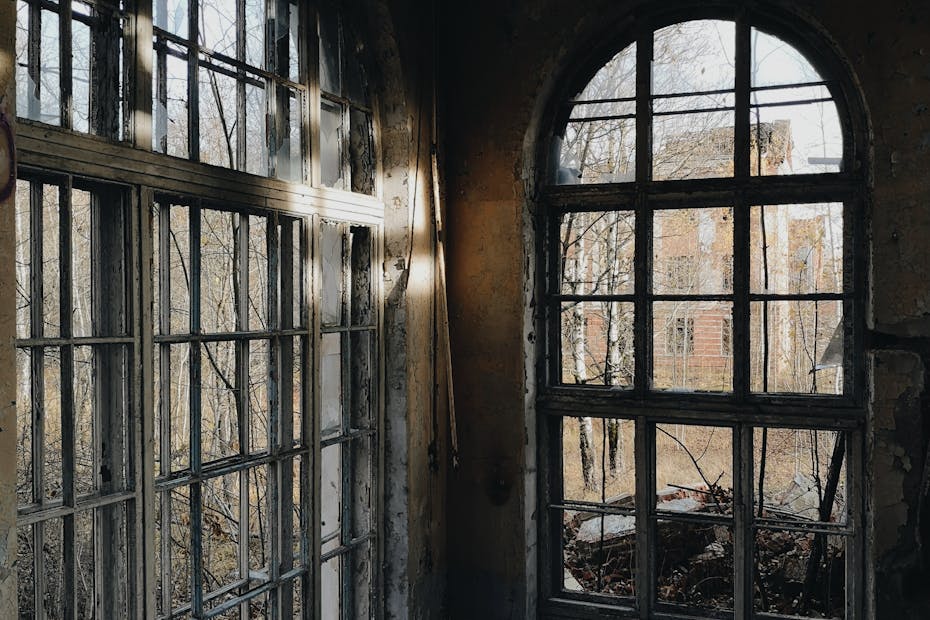 Interior of an abandoned building with large broken windows and sunlight streaming through.