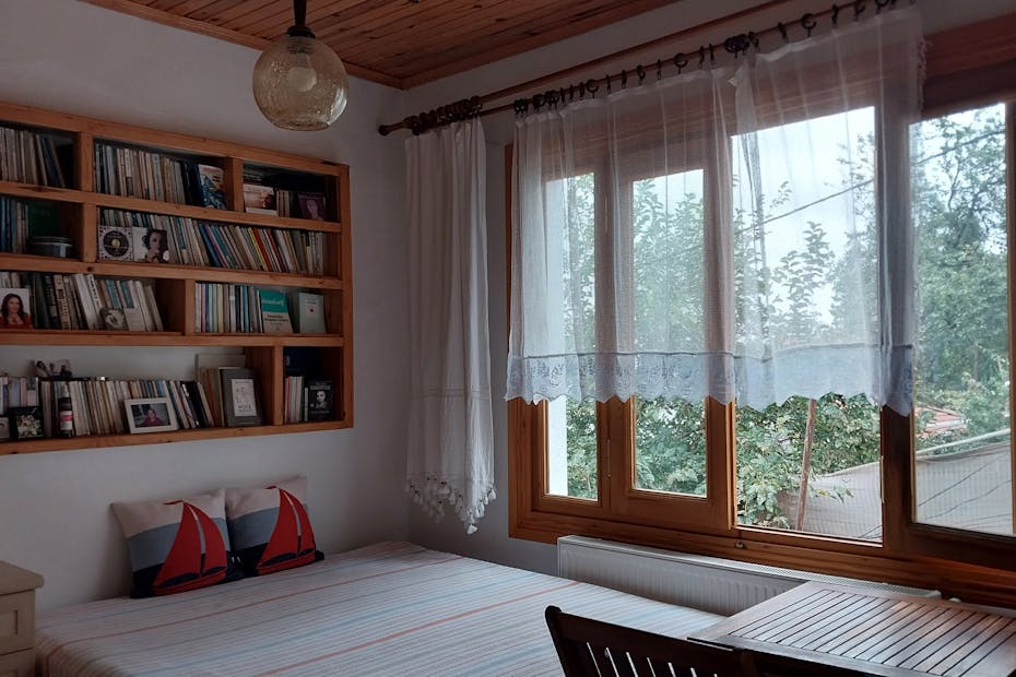 A cozy bedroom with a wooden bookshelf and a large window with sheer curtains.