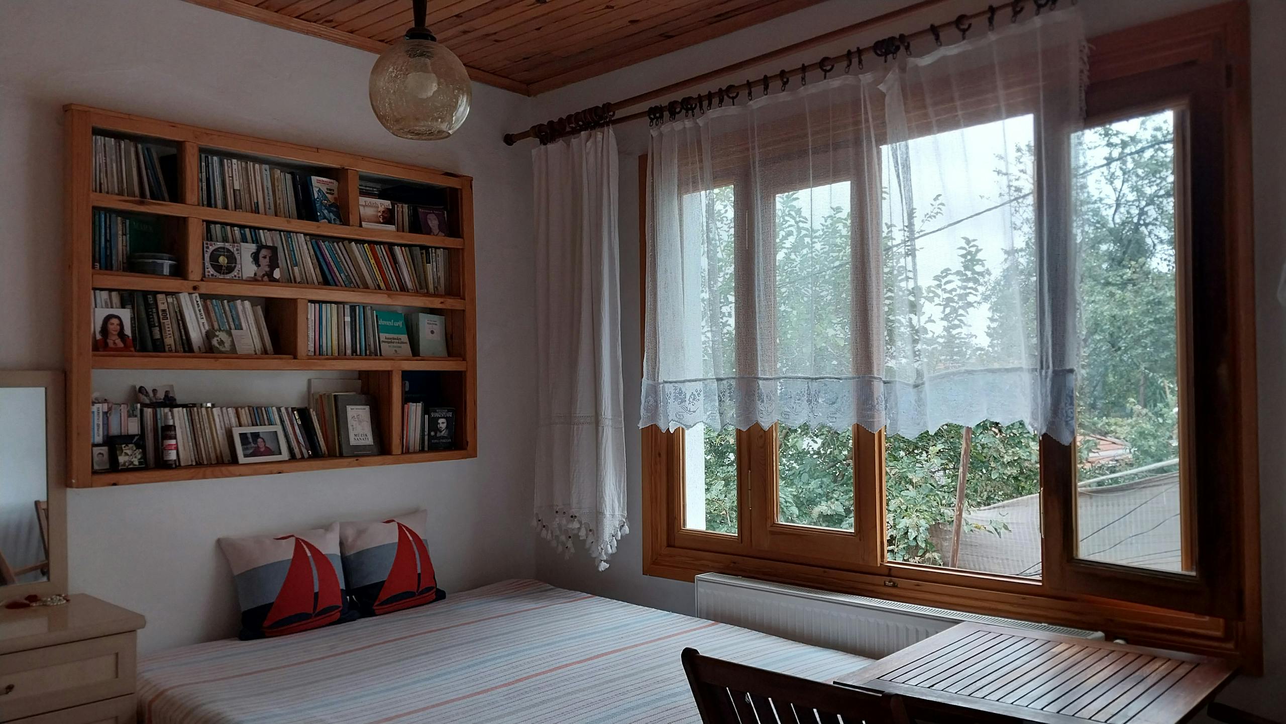 A cozy bedroom with a wooden bookshelf and a large window with sheer curtains.
