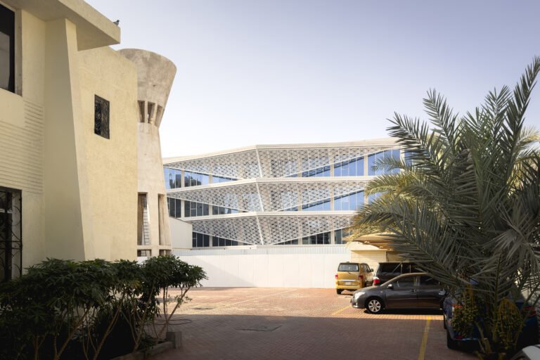 Real-life exterior architectural photograph titled 03_main facade.jpg capturing the completed main facade of the Indonesian Consulate General in Jeddah, featuring the white parametric Batik-Rowshan screen envelope, an adjacent concrete water tower, and a paved parking forecourt