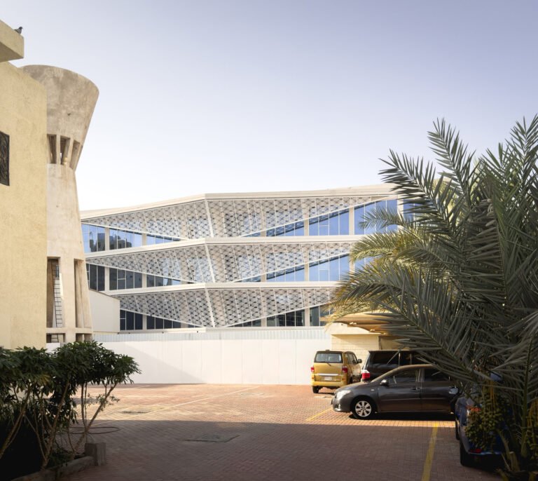 Real-life exterior architectural photograph titled 03_main facade.jpg capturing the completed main facade of the Indonesian Consulate General in Jeddah, featuring the white parametric Batik-Rowshan screen envelope, an adjacent concrete water tower, and a paved parking forecourt