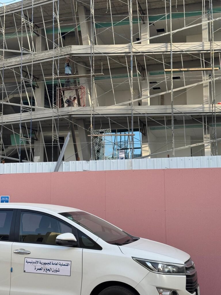 INJ Architects daytime view of a multistory concrete building enveloped in metal scaffolding rising behind a pink barrier and a parked white consulate vehicle.