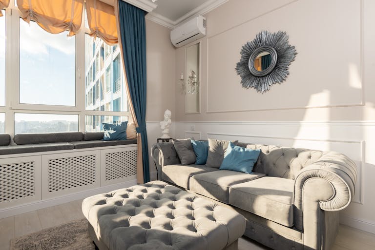 Soft couch with cushions near pouf placed at wall with mirror near big window in modern living room with bright sunlight