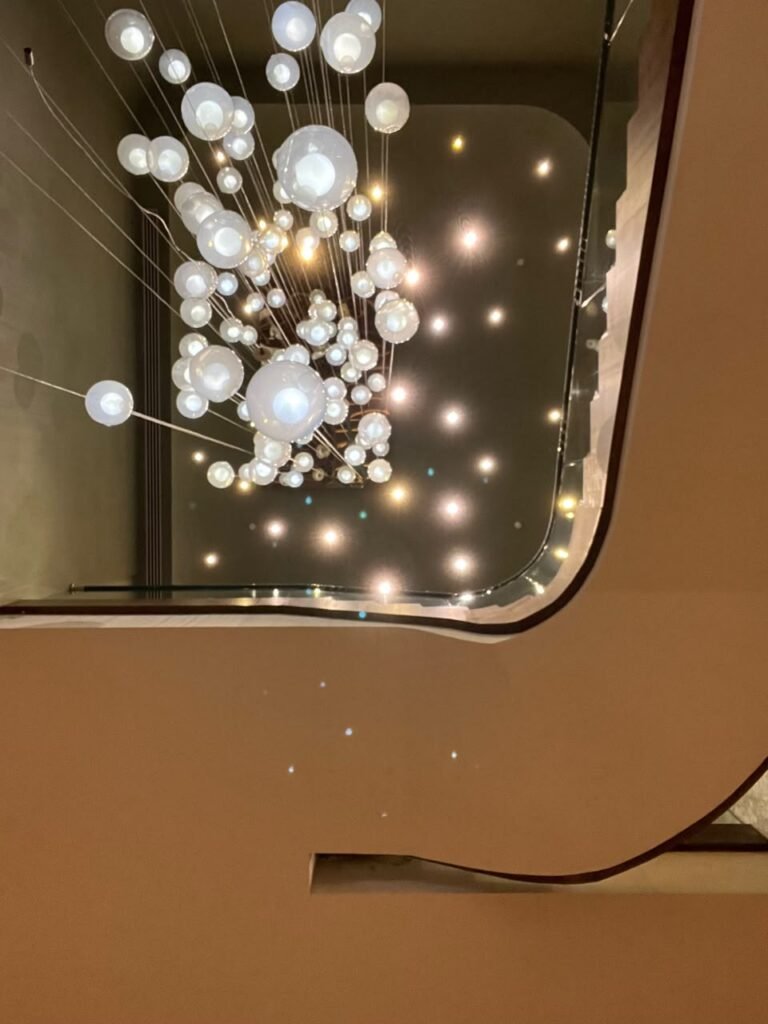 Orthographic ceiling documentation of a hotel lobby atrium, featuring a cascading chandelier of illuminated glass spheres, a curved staircase opening, and scattered recessed pinspot lighting.