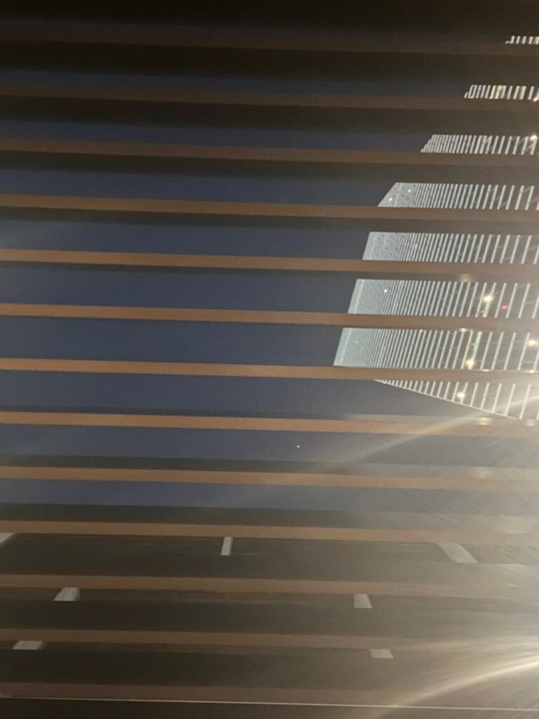 Orthographic ceiling documentation of an open-air structure featuring a rhythmic sequence of horizontal timber slats overlooking a brightly lit modern skyscraper against a dark night sky.