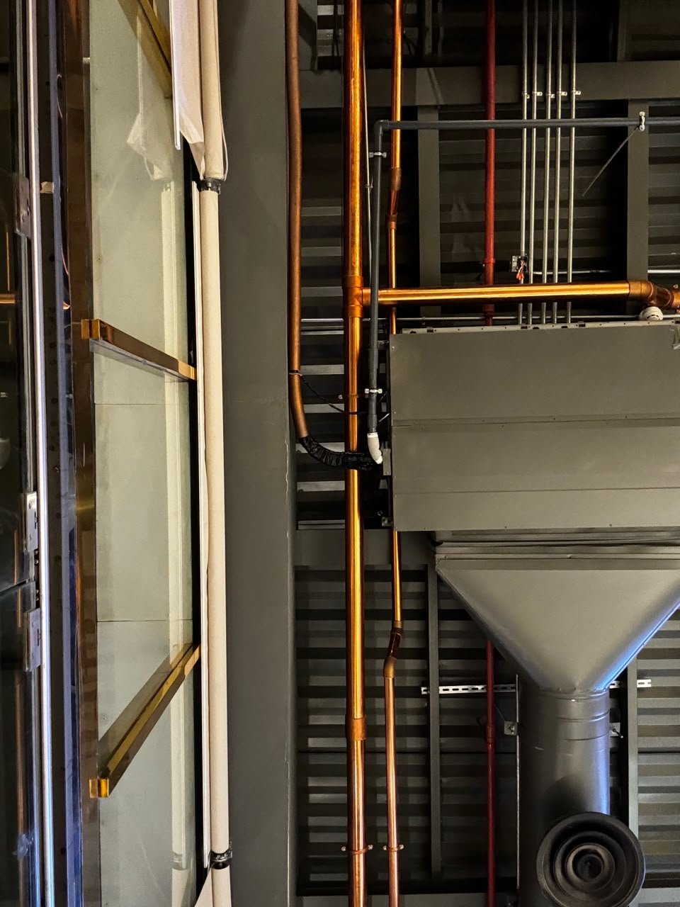 Orthographic ceiling documentation of a cafe in Taif, Saudi Arabia, showcasing an industrial open plenum with polished copper piping, a grey HVAC duct assembly with a circular diffuser, and parallel electrical conduits against a dark ribbed deck next to a brass-framed glass structure.