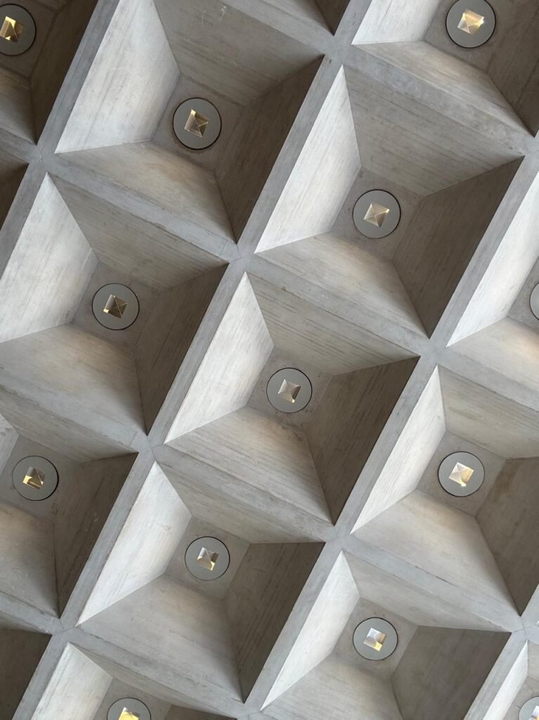 Orthographic bottom-up view of a board-formed cast-in-place concrete coffered ceiling at the Louvre in Paris, featuring deeply recessed pyramidal coffers with central circular recessed spotlights and visible timber grain textures.
