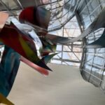 Orthographic low-angle view looking up at the complex structural glass roof canopy and steel framework of the Fondation Louis Vuitton in Paris, framed by a curved white wall and a suspended polychromatic sculptural installation.
