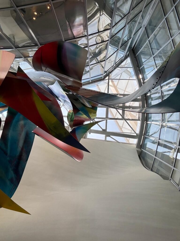Orthographic low-angle view looking up at the complex structural glass roof canopy and steel framework of the Fondation Louis Vuitton in Paris, framed by a curved white wall and a suspended polychromatic sculptural installation.