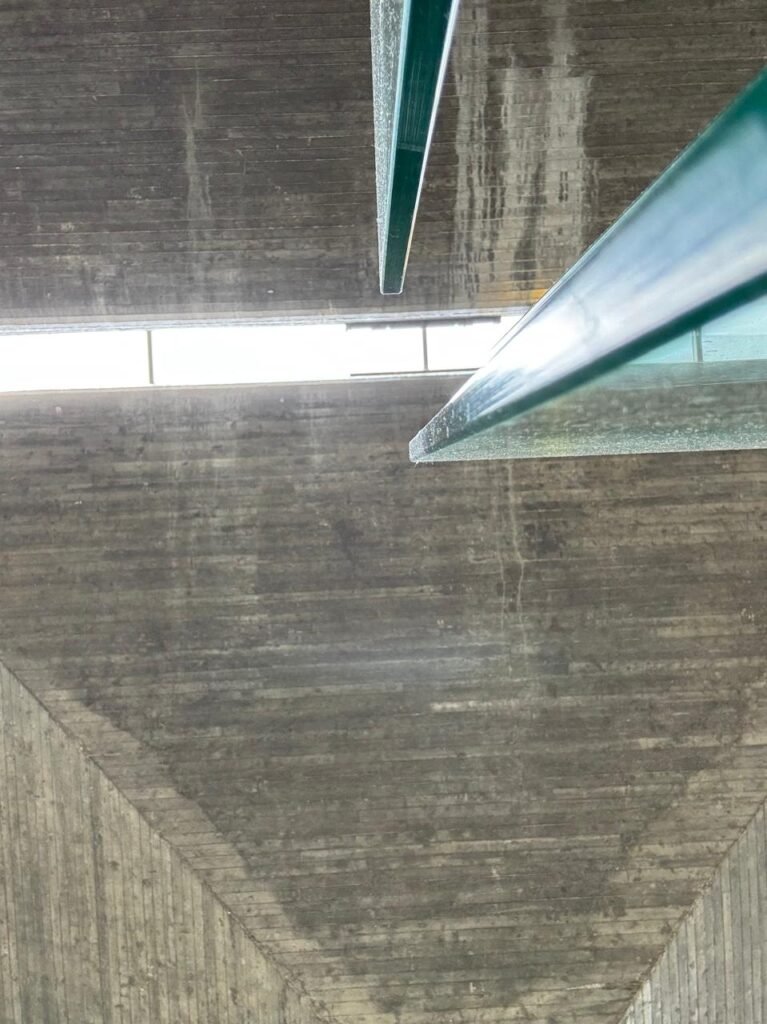 Orthographic bottom-up architectural view of a board-formed exposed concrete soffit at a Charles de Gaulle Airport transit station, displaying wood-grain texture impressions, a linear ceiling reveal gap, and cantilevered structural glass fins.