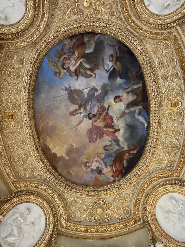 Orthographic bottom-up view looking at a historic French Classical ceiling at the Louvre Museum in Paris, displaying a central oval allegorical fresco painting framed by rich gilded stucco moldings, intricate foliage relief carving, and corner white plaster bas-relief medallions.