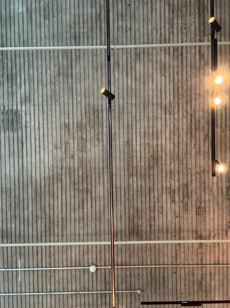 Orthographic view looking up at a linear board-marked exposed concrete ceiling soffit with black suspended lighting tracks, directional spotlights, and white horizontal conduits.