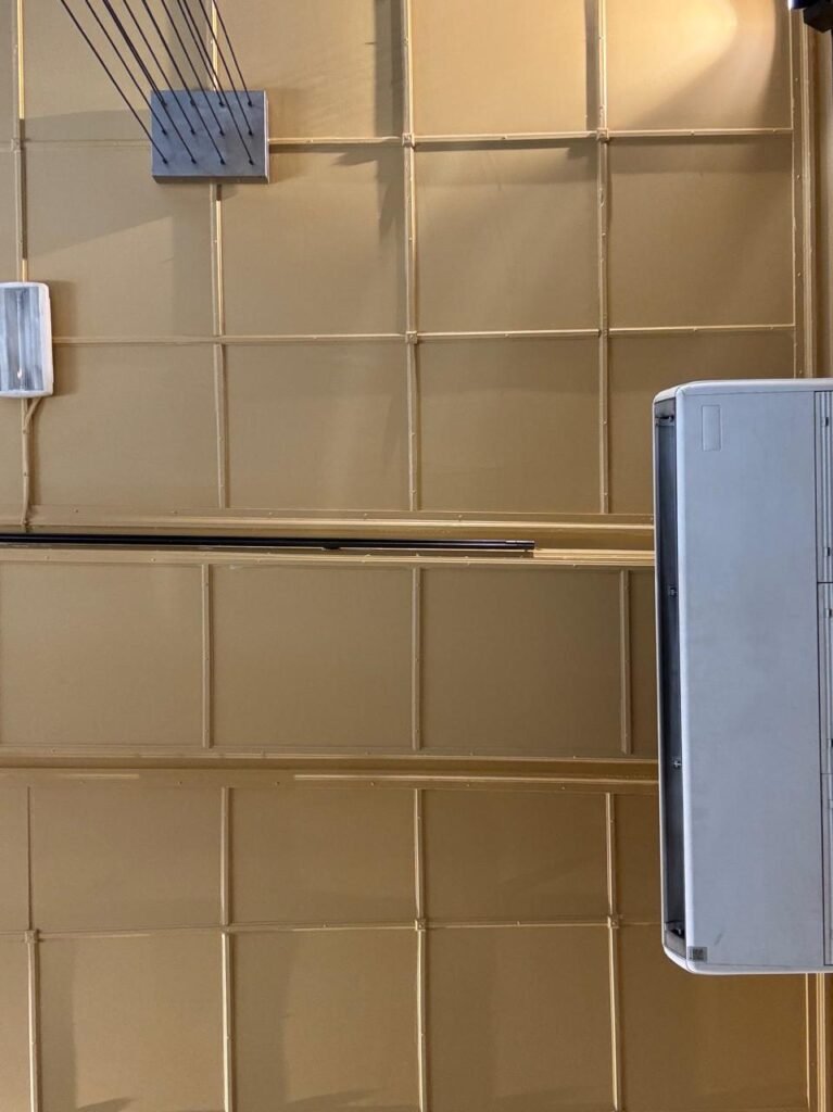 Low-angle orthographic photograph of a gold-painted coffered metal grid ceiling with surface-mounted conduits, pendant suspension wires, a fluorescent fixture, and a white split-system AC unit.