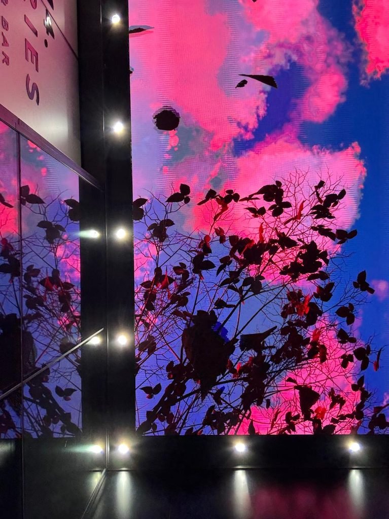 Low-angle view of a digital LED ceiling screen displaying a hyper-saturated pink and blue sky with tree branch silhouettes, flanked by vertical black frame members with glowing warm LED bulbs.