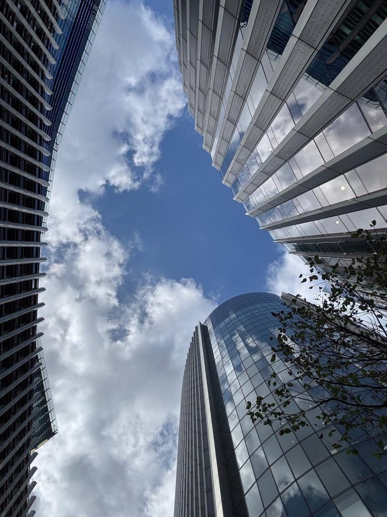 Low-angle orthographic photograph looking directly upward into an open-air skyscraper plaza framed by curved glass and steel high-rise facades against a blue sky with white clouds.