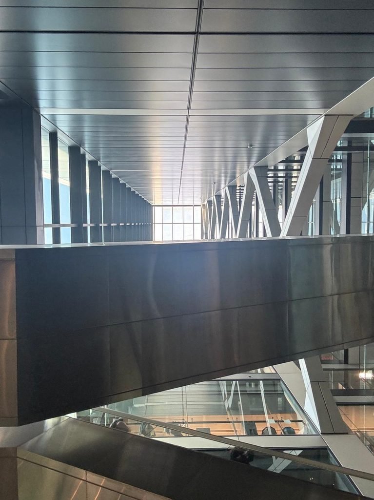 Interior architectural view looking up through a multi-story skyscraper atrium, featuring metallic modular ceiling panels, massive white diagonal structural steel V-braces along glazed curtain walls, and cantilevered stainless steel skybridges with glass balustrades.