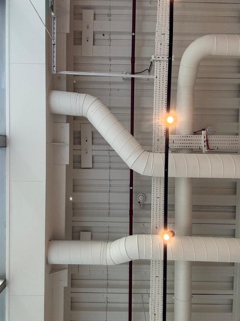 Orthographic view looking up at an industrial exposed ceiling with white-painted ribbed metal decking, thick insulated white HVAC pipes with curved elbows, perforated cable trays, a thin red fire pipe, and a suspended black light track with warm spotlight bulbs.