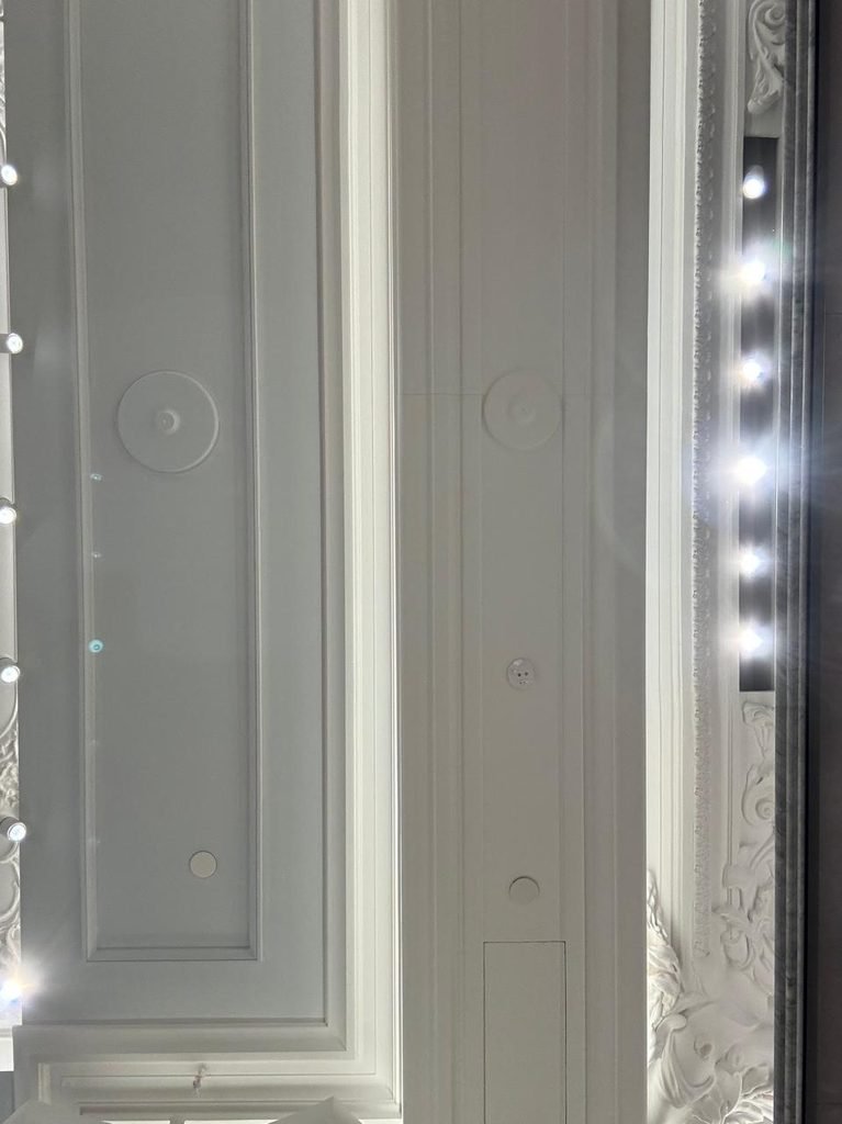 Classic white ornamental plaster ceiling at Harrods London featuring stepped linear moldings, floral ceiling roses, intricate relief details, and a side lighting track with illuminated bulb fittings.