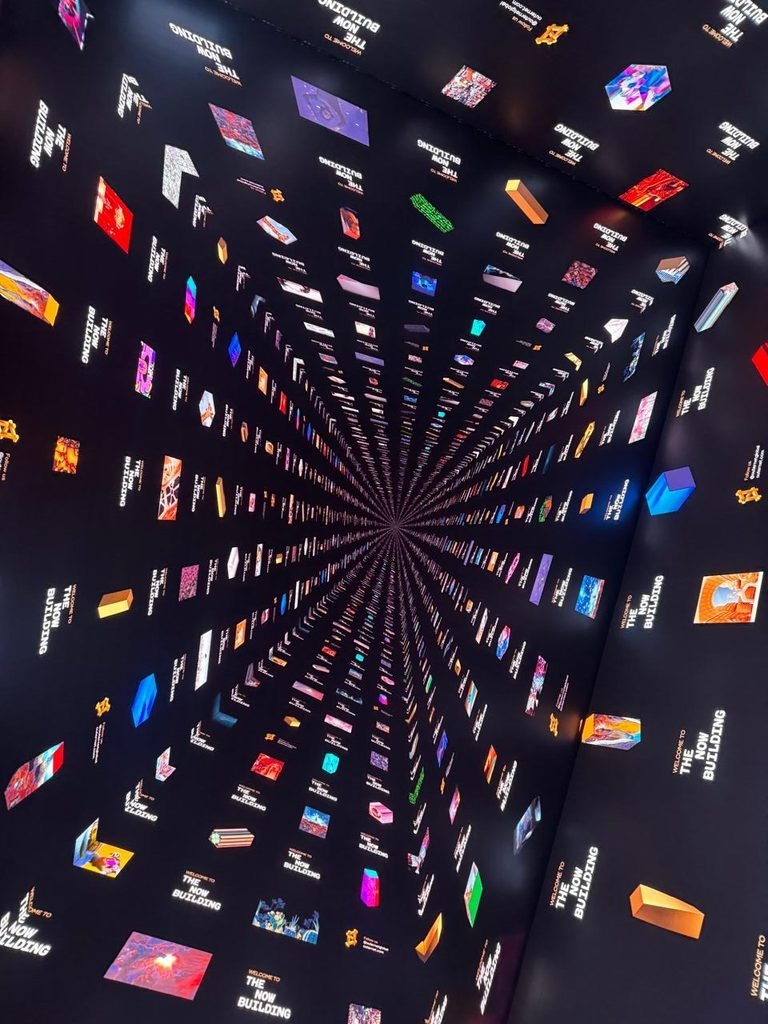 A giant LED digital ceiling and wall display inside a modern immersive hall, showcasing an optical illusion 3D pattern of digital cubes and media extending toward a center vanishing point with "The Now Building" typography.