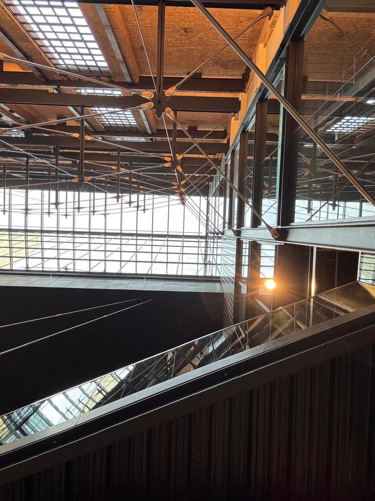 Low-angle perspective looking up at a dramatic industrial glass skylight roof supported by steel tie-rod trusses, brick coffered ceiling arches, and glass-clad escalator walkways.