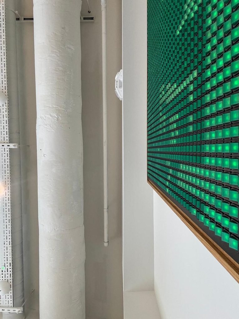 Orthographic side view of white vertical building service pipes and perforated cable trays running along a wall, adjacent to a glowing green LED matrix pegboard display screen.