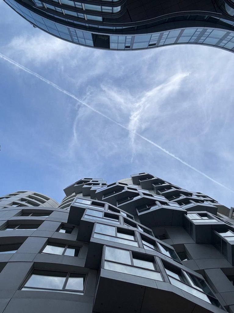 Skyward zenith photograph looking directly up at the sculptural, undulating glass facade of Frank Gehry's Prospect Place buildings framing a clear blue sky with contrails.