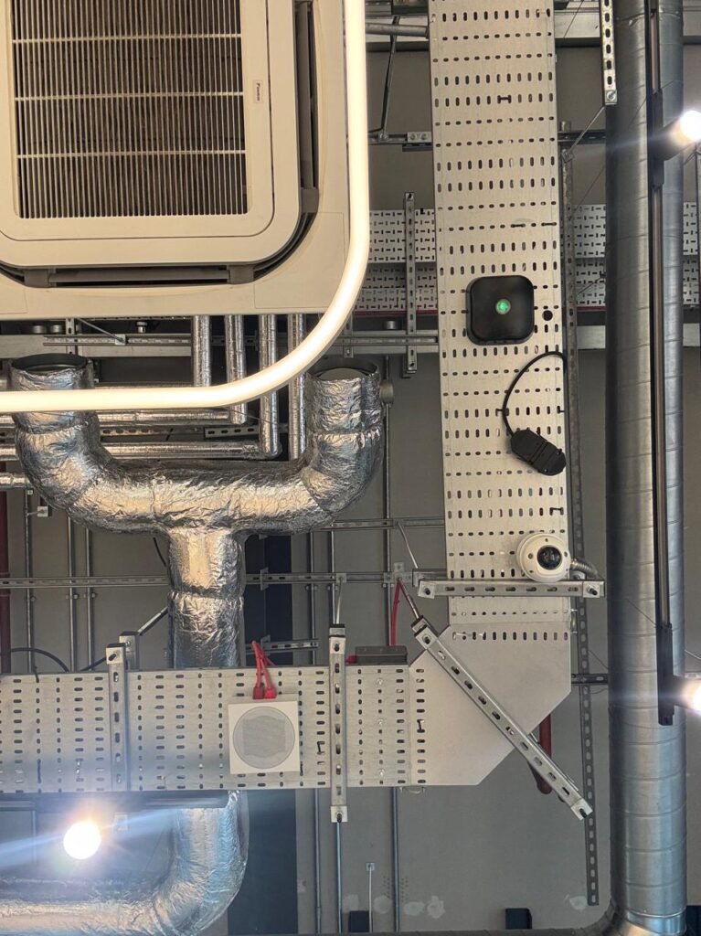 Low-angle orthographic photograph of a complex open-soffit MEP ceiling featuring a square AC cassette unit wrapped in a glowing halo LED ring, silver foil Y-ducts, perforated tray raceways, and a dome camera.
