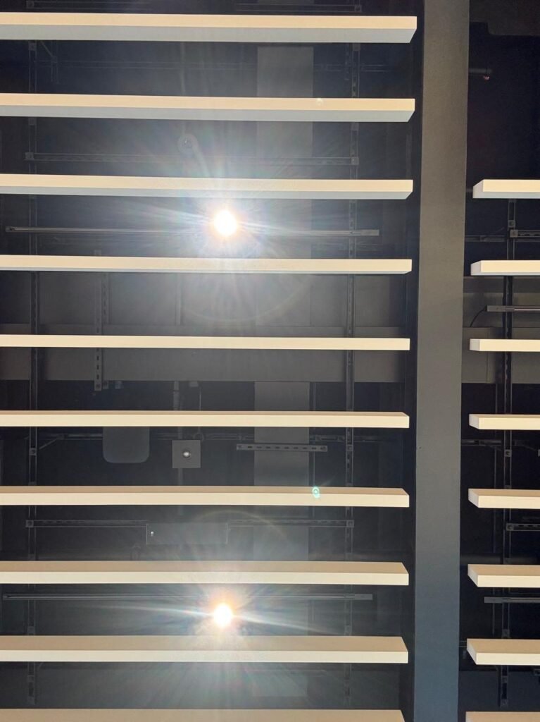 Orthographic low-angle view of white horizontal acoustic ceiling baffles suspended below a dark painted soffit, featuring bright downlight lens flares and a solid dark structural column on the right.