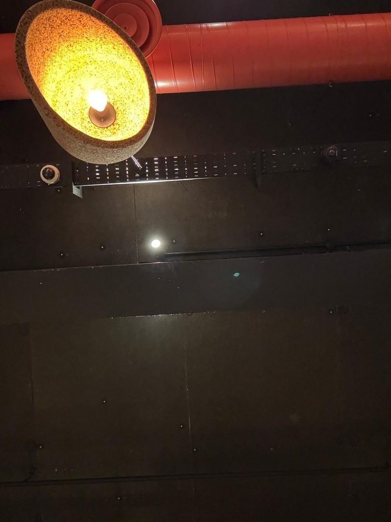 Orthographic view looking up at a dark painted ceiling soffit featuring a thick orange duct, a textured curved pendant shade emitting warm light, and a black perforated cable tray equipped with security sensors.