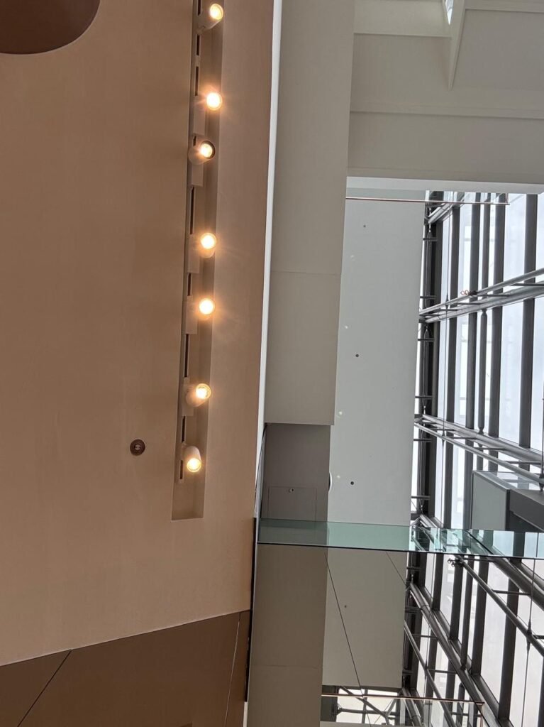 Orthographic view looking up at a multi-level atrium soffit with a linear strip of warm globe light bulbs, a glass balustrade edge, and a high structural steel and glass curtain wall.