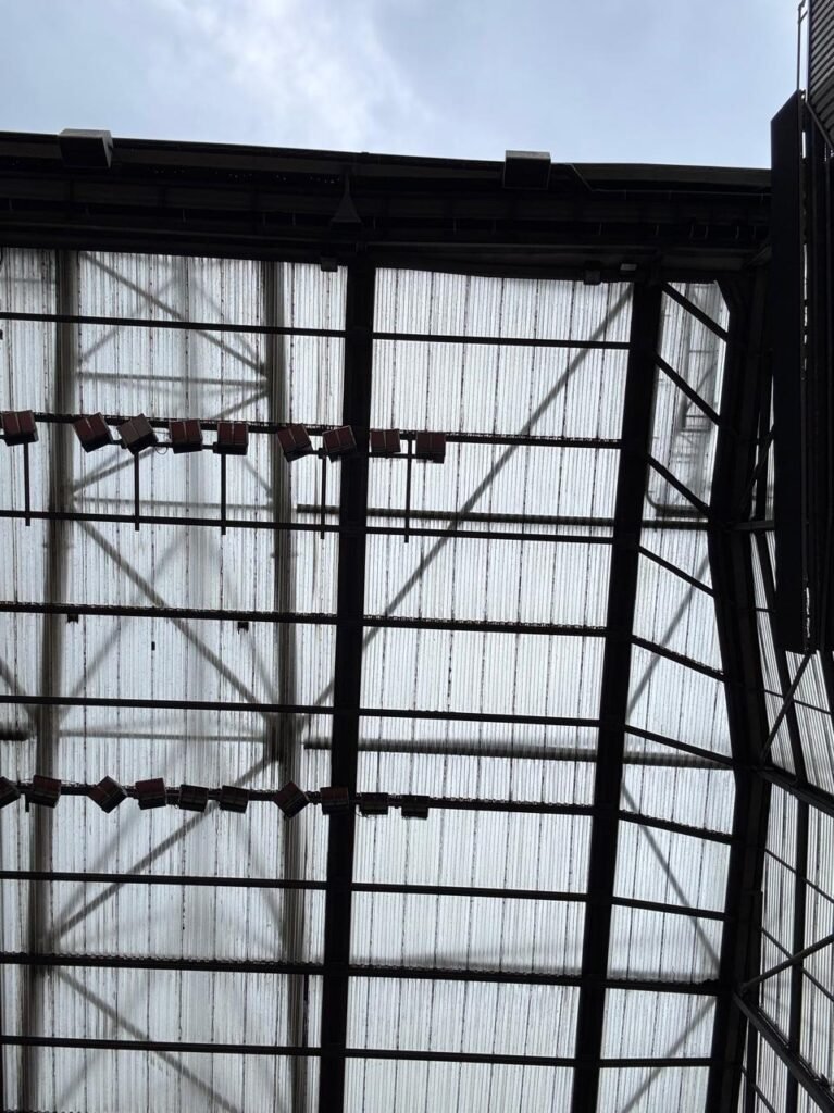 Orthographic low-angle photograph looking up at Old Trafford's cantilevered stadium roof structure, featuring translucent corrugated cladding panels, heavy black steel trusses, and suspended floodlight arrays.