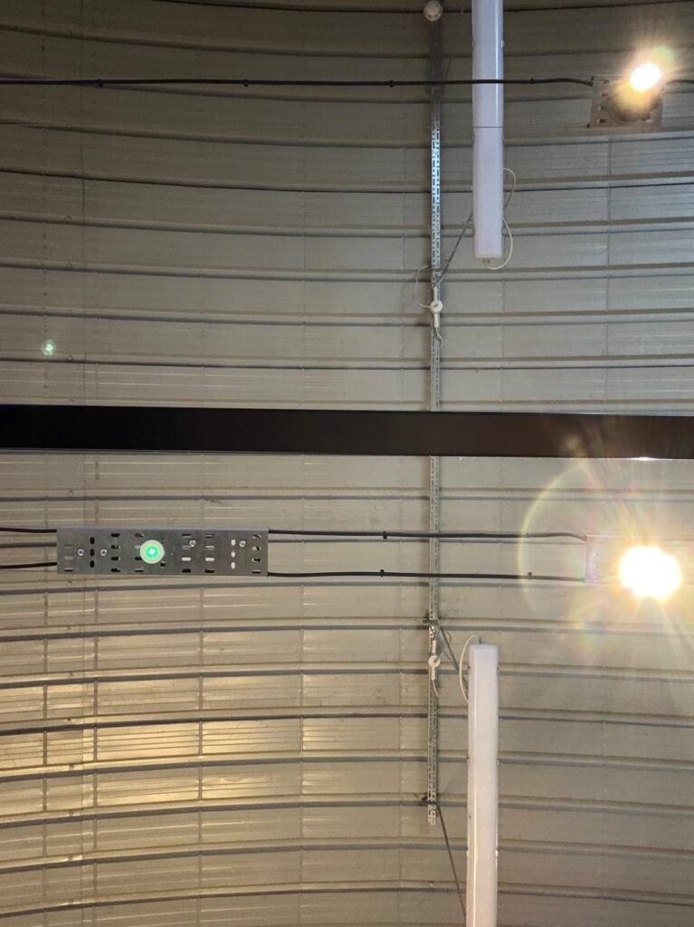 Orthographic view looking up at a curved corrugated metal barrel-vault ceiling soffit in light grey, equipped with black cable runs, a central black beam, suspended linear tube lights, and perforated galvanized cable raceways with indicator lights.
