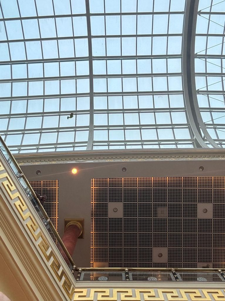 Low-angle orthographic photograph looking up at a grand glazed barrel-vault skylight roof with white steel ribs, supported above a classical interior balcony with gold Greek key ornamental trim and perforated acoustic ceiling tiles.