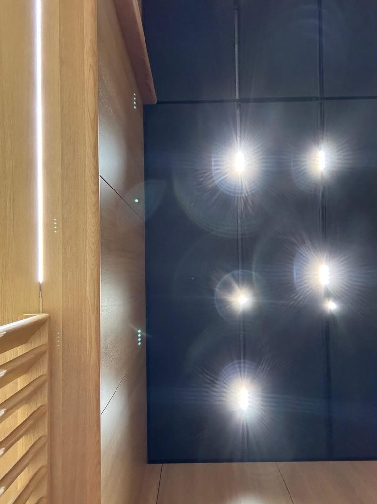 Orthographic view looking up at a matte black acoustic panel ceiling with intense warm spotlight lens flares, flanked by a natural oak wall panel featuring linear vertical LED light strips.