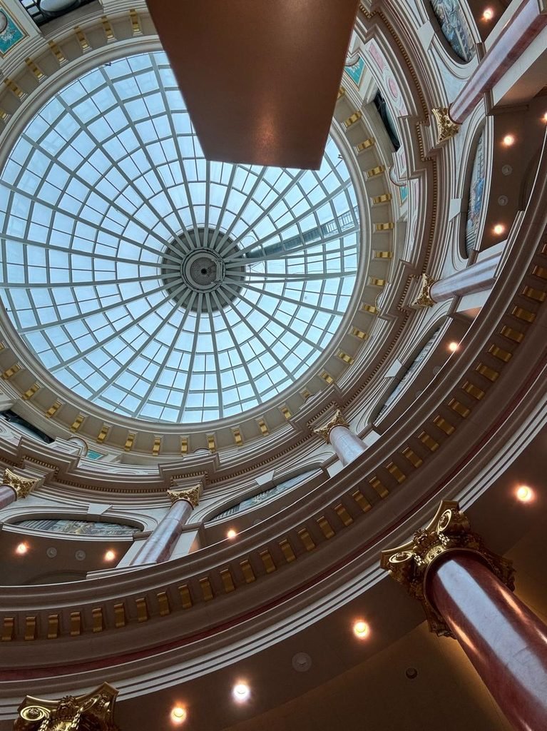Low-angle orthographic photograph of a massive Neoclassical radial glass dome skylight framed by multi-tiered circular balconies, gold Corinthian columns, and faux-marble pillars.