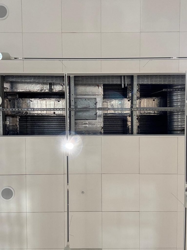 Orthographic photograph looking up at a white modular suspended ceiling with a clean rectangular reveal exposing insulated silver foil HVAC ducts, cable trays, and structural channels above.